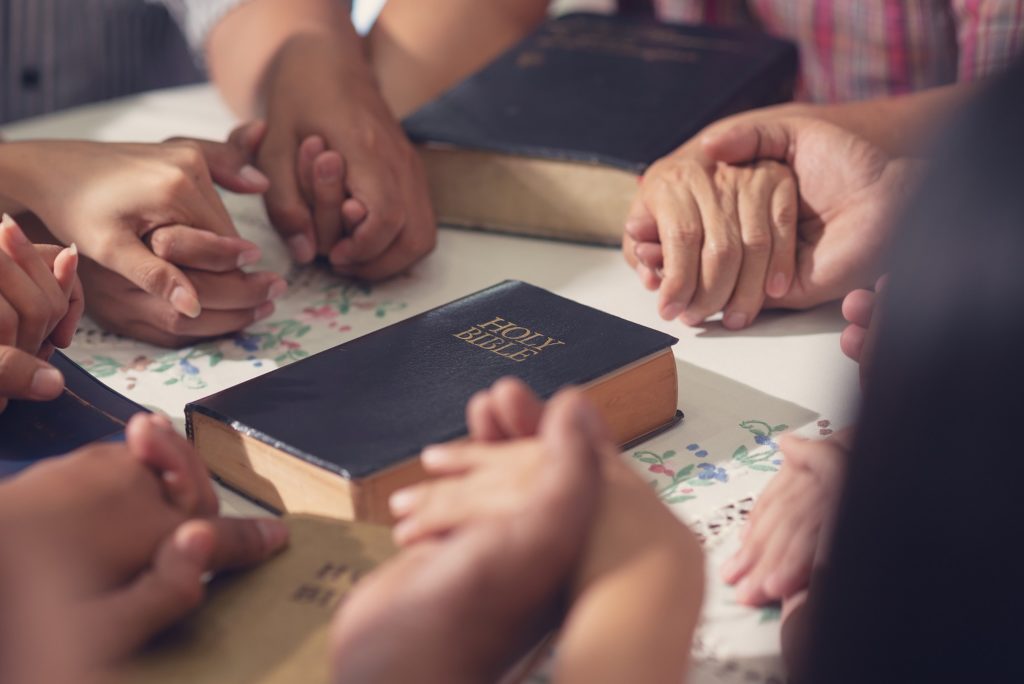 close up of Holy Bible in the middle of a prayer group (only clasped hands visible) that is fulfilling prayer requests