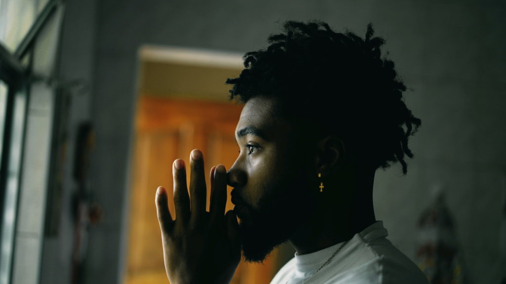 A young man holding his hands in prayer as he looks out the window, deep in thought.