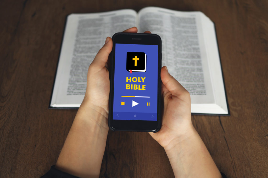 A person holding a smartphone that says “Holy Bible” on it, with a Bible in the background.